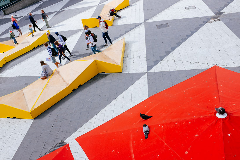 Kids and pigeons sitting at yellow seating installations at the Sergels torg Plaza in Stockholm.