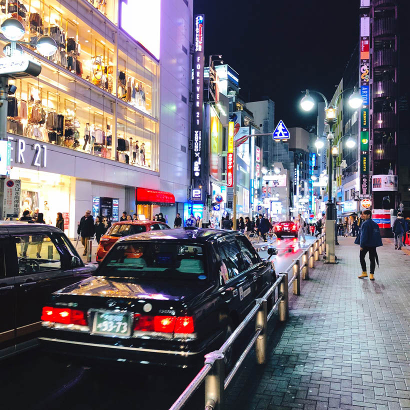 A taxi car driving around a shopping street in Shibuya while the area is being lit by the signs of the nearby buildings in Tokyo, Japan.